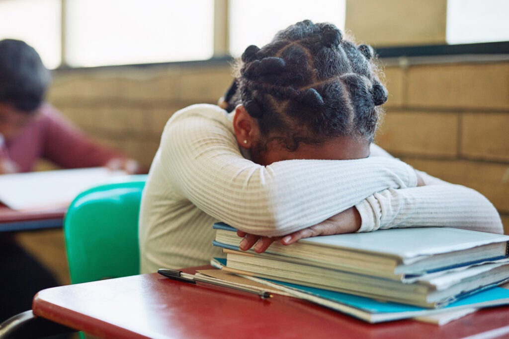 A young Black student sits at her desk with her head down on a pile of books.