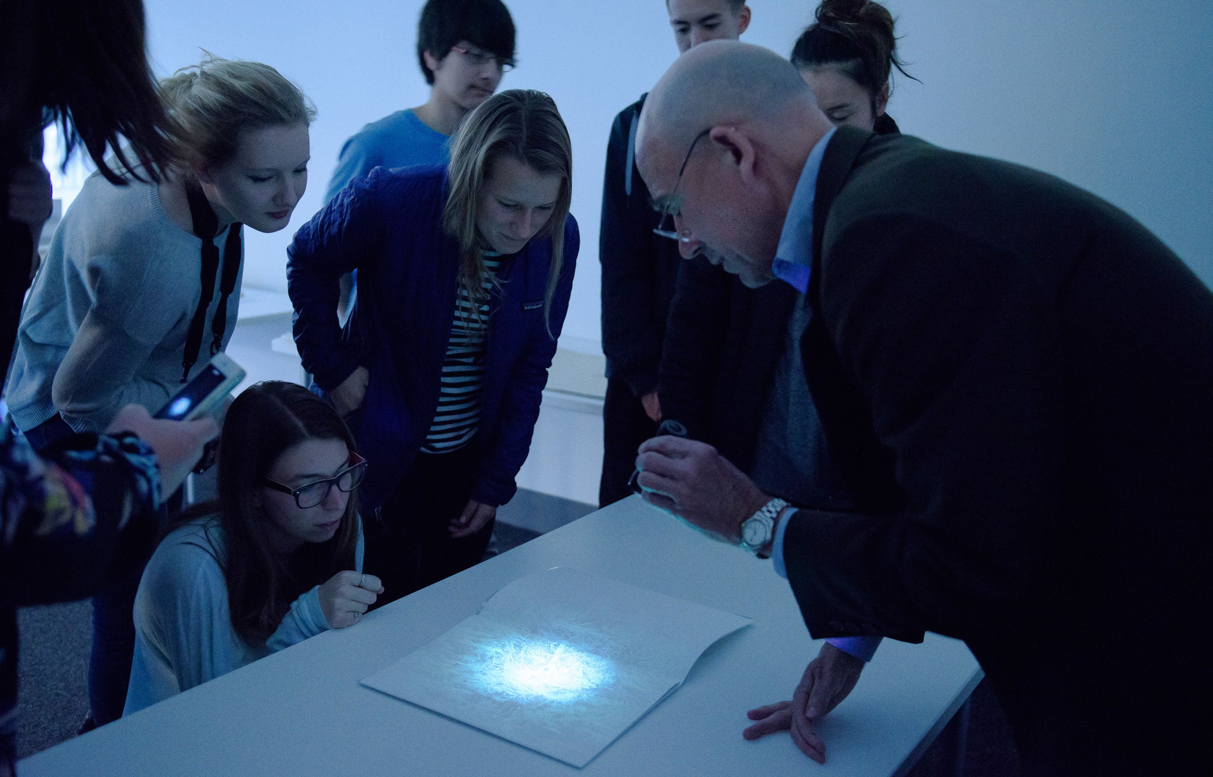 Undergraduate students, crouching, leaning and standing around a table as a male librarian dressed in a dark suit jacket and blue dress shirt focuses a beam of light from a very small UV flashlight on a page in 2013 (Image credit: L.A. Cicero)