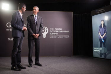 Sundar Pichai and Barack Obama speaking with a screen showing a young woman in the background.