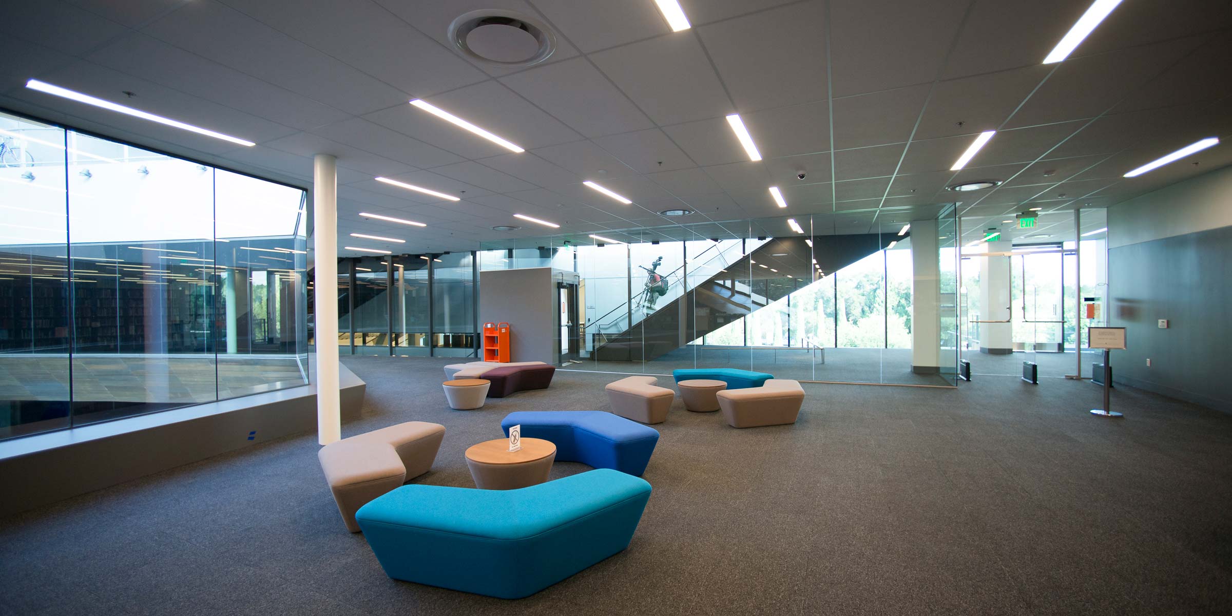 Three groupings of upholstered benches in turquoise, blue, beige and brown stand next to round end tables in an open space near the library's main desk, with glass walls and windows in the background.) (Image credit: L.A. Cicero)