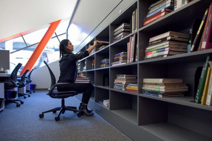 Alexandria Brown-Hedjazi, an art history graduate student, dressed in black, sits on an office chair in her carrel, a pile of books in her lap. She leans forward to lift a book from her bookcase. (Image Credit: L.A. Cicero)