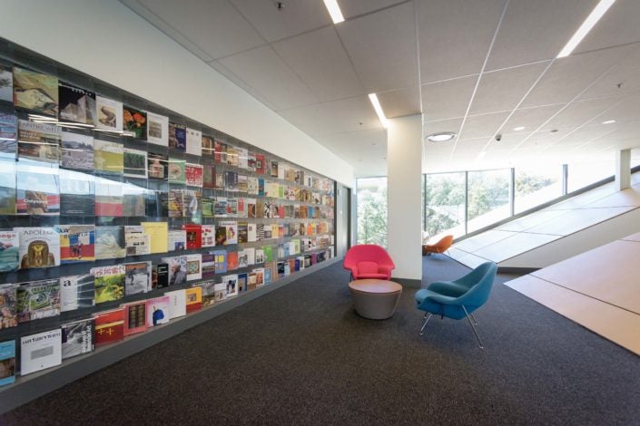 A colorful wall of periodicals on the left, with an entryway set in a steeply sloped wall on the right. In between are upholstered armchairs – one tangerine, another turquoise – with a small, low round table in between. (Image Credit: L.A. Cicero)