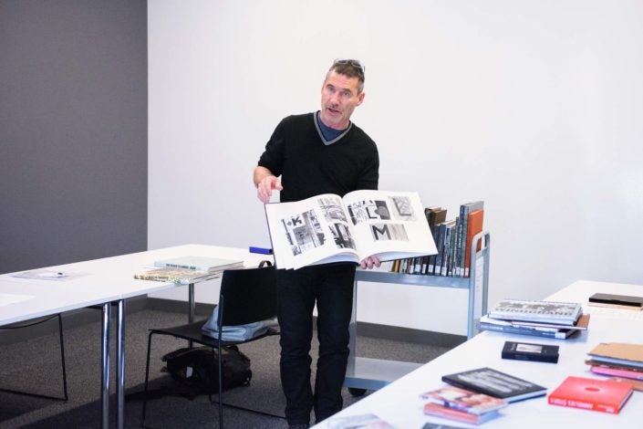 Professor Lukas Felzmann dressed in black, with eyeglasses on the top of his head, stands between two tables in a classroom filled with natural light, pausing in the middle of turning the pages of a large photographic book. One of the tables is strewn with a dozen books and a metal cart behind him holds a dozen more. (Image Credit: L.A. Cicero)
