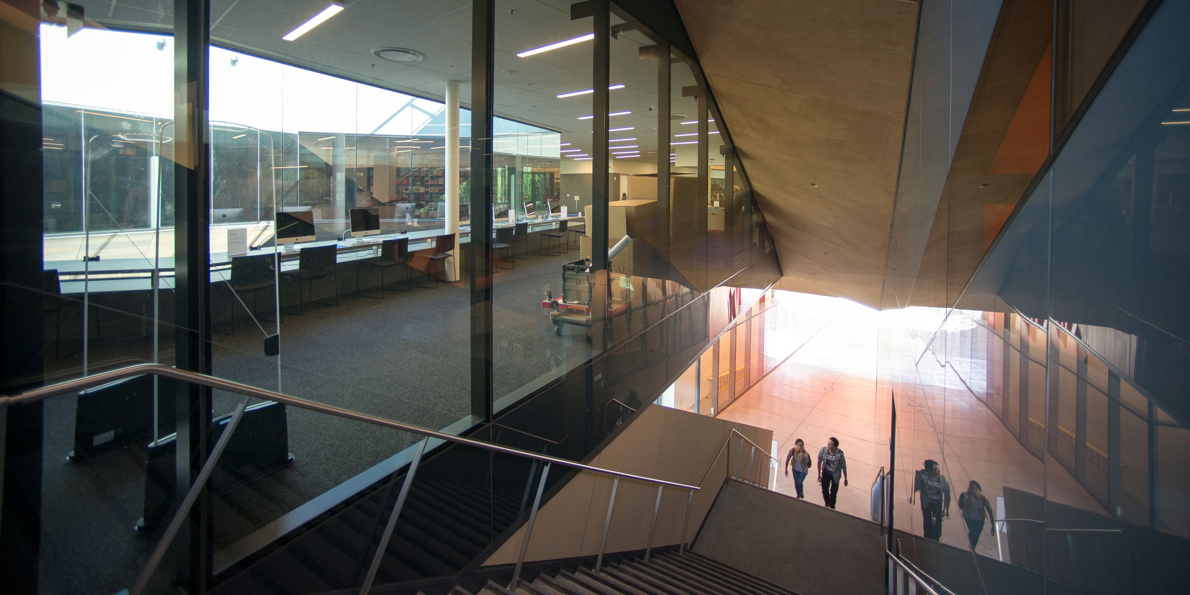 Two people ascending the covered staircase that leads to the library from the McMurtry Building's courtyard. The photo shows the floor-to-ceiling glass windows of the library, with a view of a long table that frames one side of the oculus. On the table are six desktop computers. (Image credit: L.A. Cicero)
