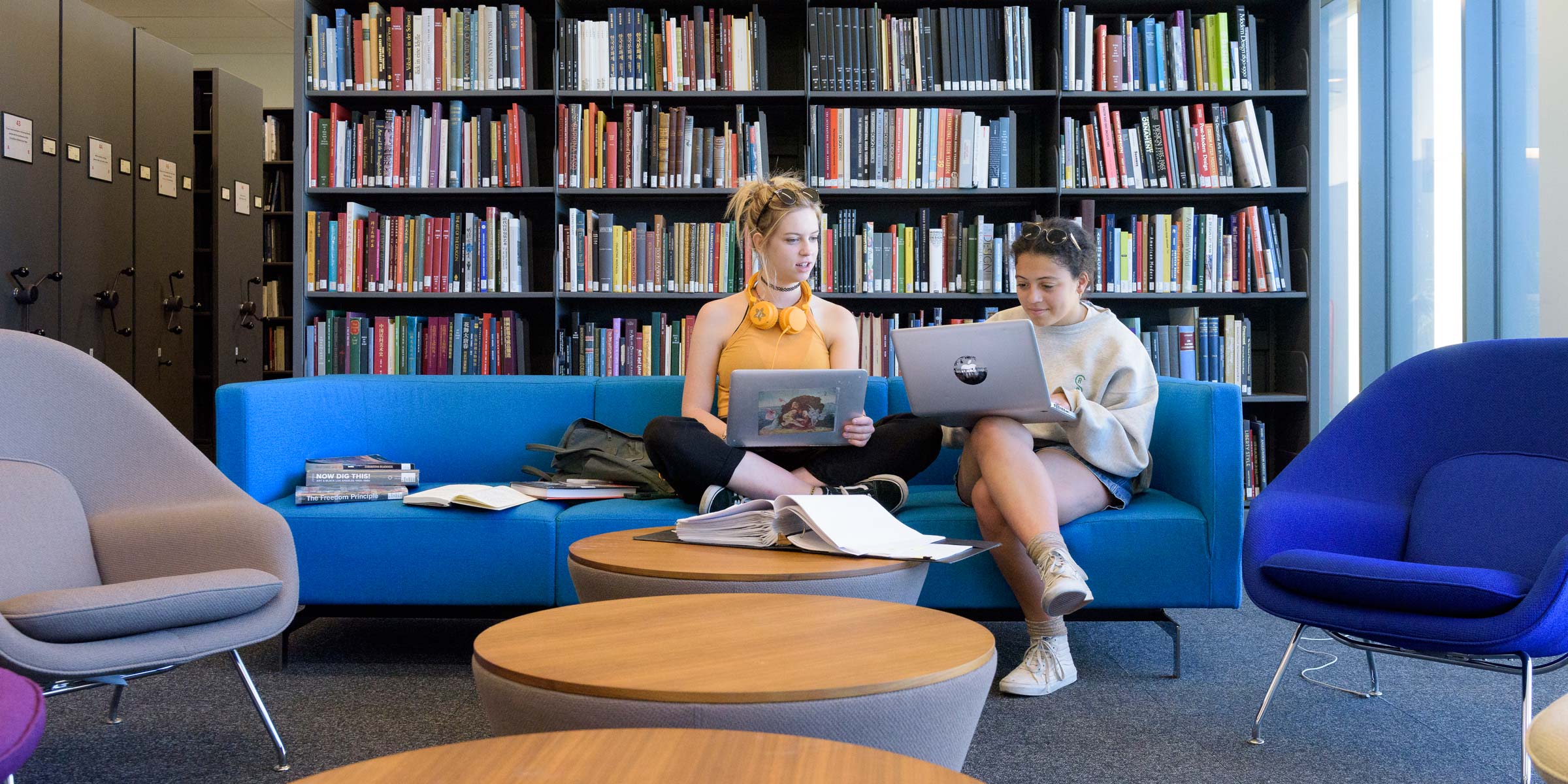 Juniors Clara Galperin and Josie Hodson, sunglasses on the tops of their heads, sit close together at one end of a bright blue couch with open laptops in one section of the library's open stacks. Behind them is a tall bookcase filled with books (Image credit: L.A. Cicero)