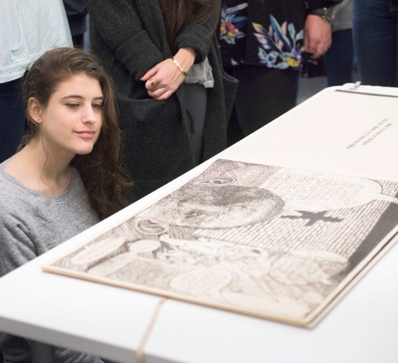 Jacqueline Langelier, an undergraduate student with long brown hair kneels on the floor while studying a page in Cry Uncle. The rest of the class stands behind her looking over her head. (Image Credit: L.A. Cicero)