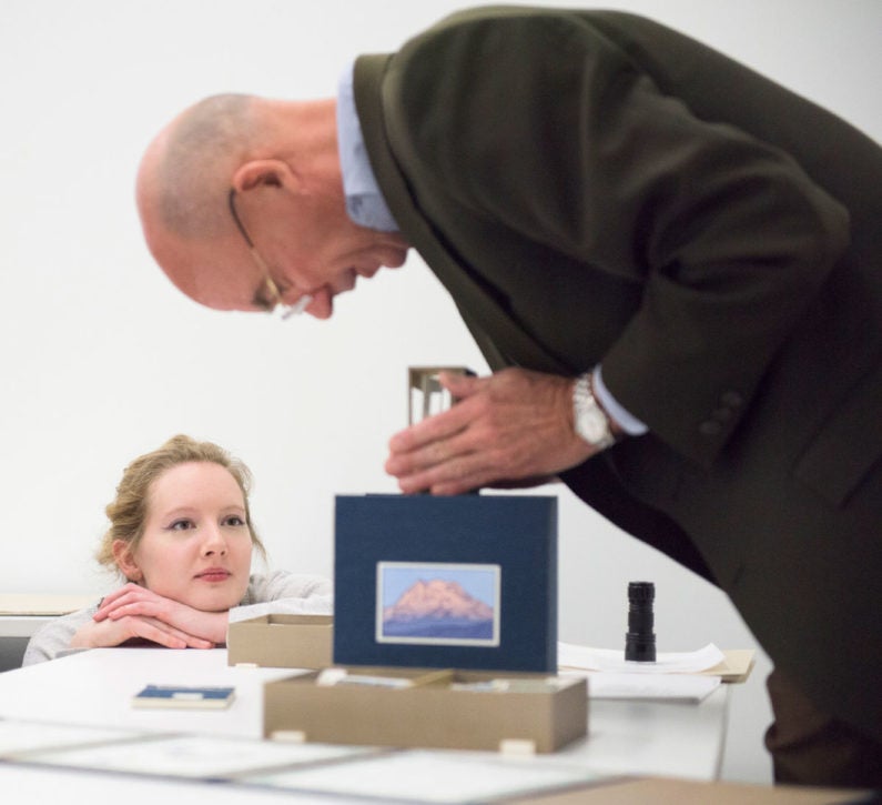 Hadley Nelson, an undergraduate student with blond hair kneels on the floor, her chin resting on her hands on top of the table, as she watches Peter Blank, dressed in a dark suit and blue shirt bending a 10-inch cube covered in dark blue silk with a hand-colored image of Mount Rainier. It is an artist's book titled Local Conditions: One Hundred Views of Mount Rainer – At Least. (Image Credit: L.A. Cicero)