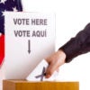 Hand inserting a ballot to voting box. The sign above the slot in two languages, English and Spanish. USA flag and white background.