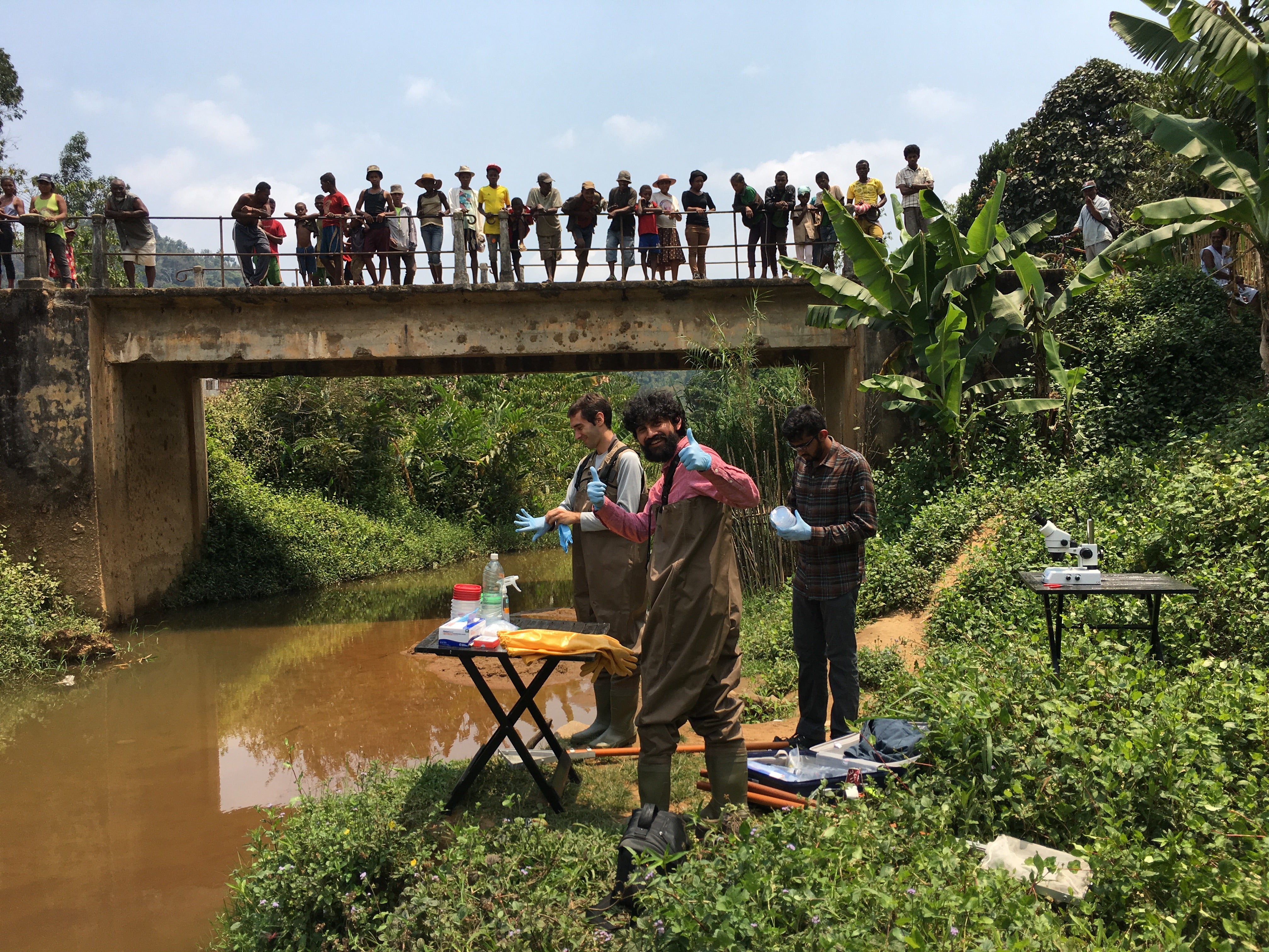 Stanford scholars Manu Prakash and Deepak Krishnamurthy with local worker Andres (no last name given) in a river in Madagascar gathering larvae that cause schistosomiasis while local people look down on them from a bridge