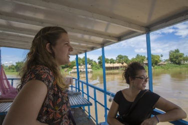 two young woman seated on a boat, traveling down a muddy river