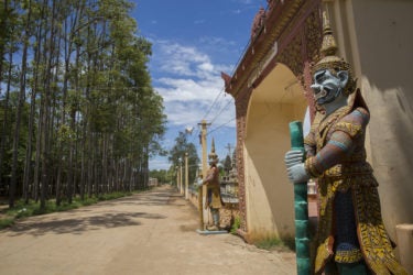 A view down a dirt road, with trees on the left, and the entrance to a temple on the right. Two ornate statues flank the temple entrance.