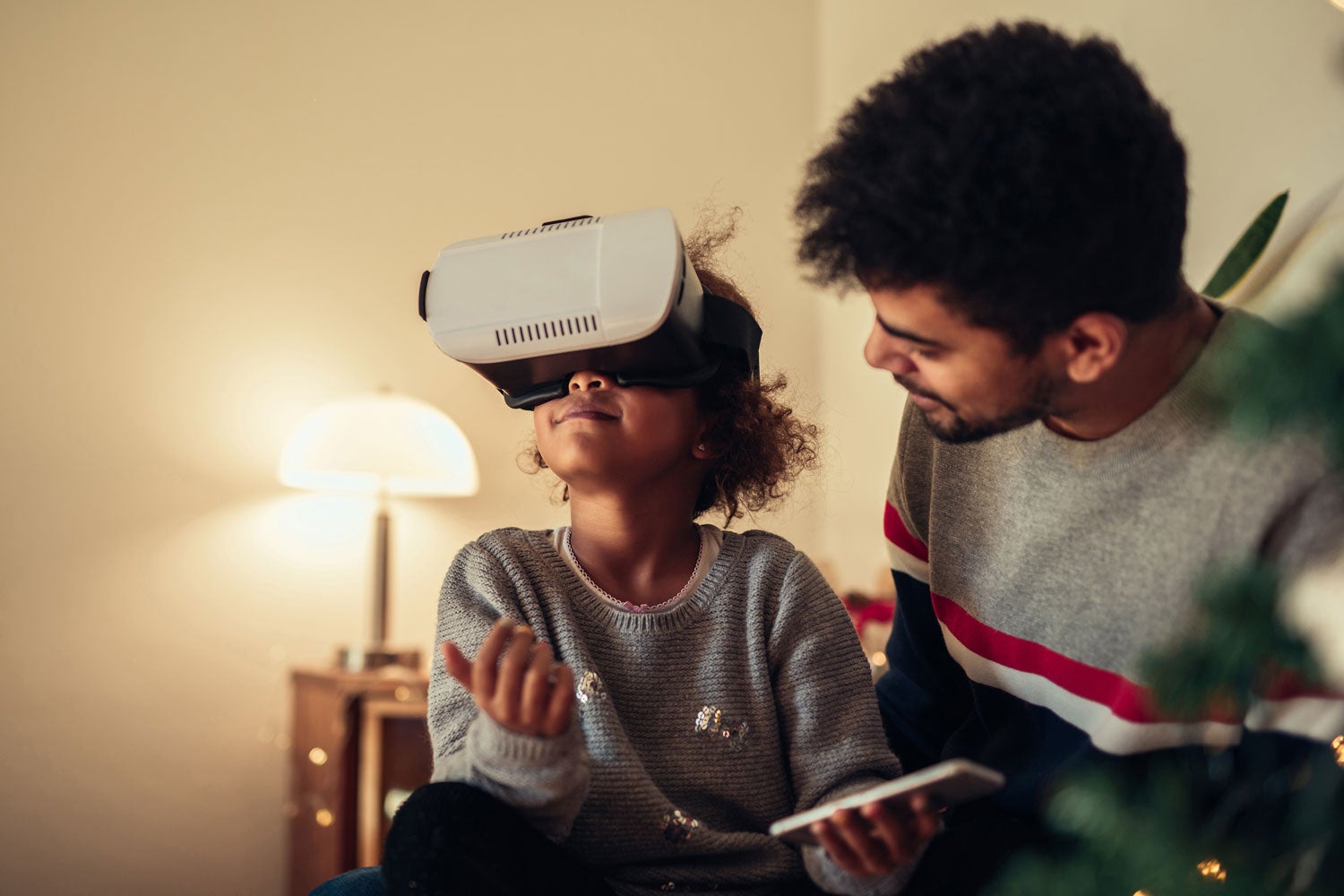 father sitting next to daughter and looking on as she wears a VR headset