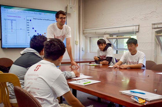 Students are rolling dice to predict the weekly spread of an infectious disease, as a way to learn about the predictive power of computational mathematics. Engineering graduate student standing is Maxime Bassenne