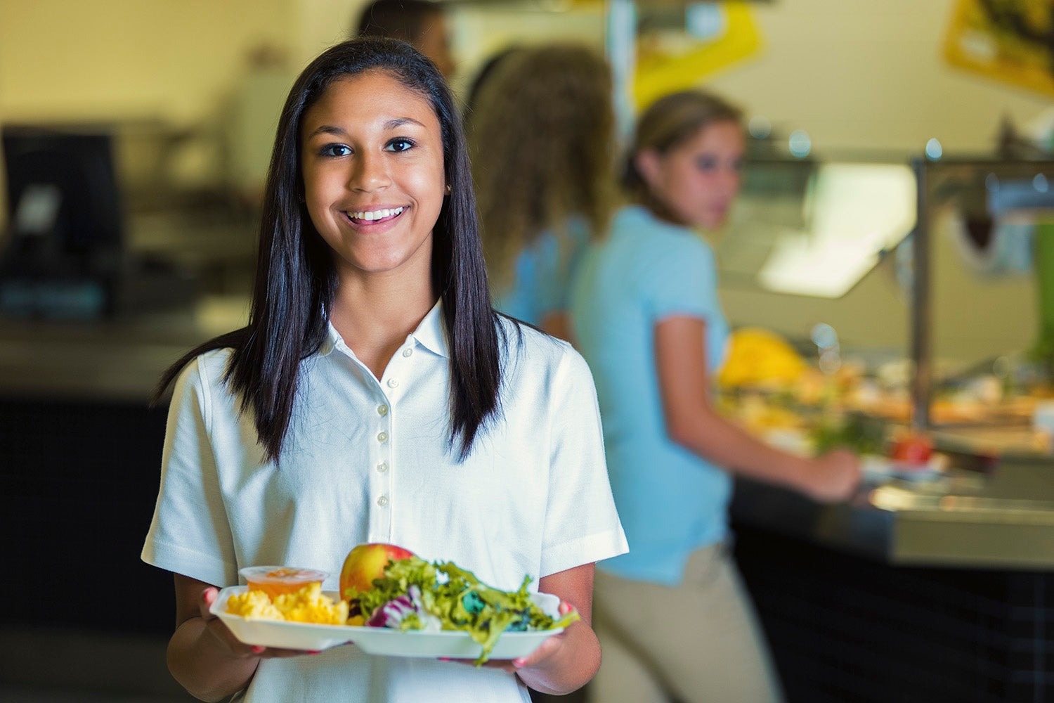 High school student holds a plate of healthy food in the school cafeteria. Her classmates are waiting in line in the background.