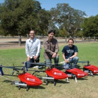 Three men kneeling behind three radio-controlled helicopters outside