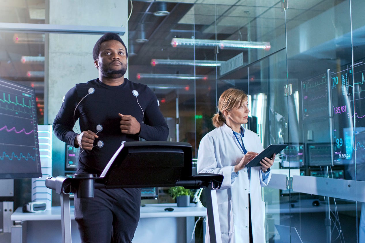 Male Athlete Walks on a Treadmill with Electrodes Attached to His Body while Sport Scientist Interacts with Touchscreen and Supervises EKG Status. In the Background Laboratory with High-Tech Equipment.