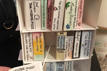 A paper bookshelf with cards made to look like books. A person is pulling on the Misérables card to show how it makes a sign reading "D-Library Favorites for Hugo" pop up.