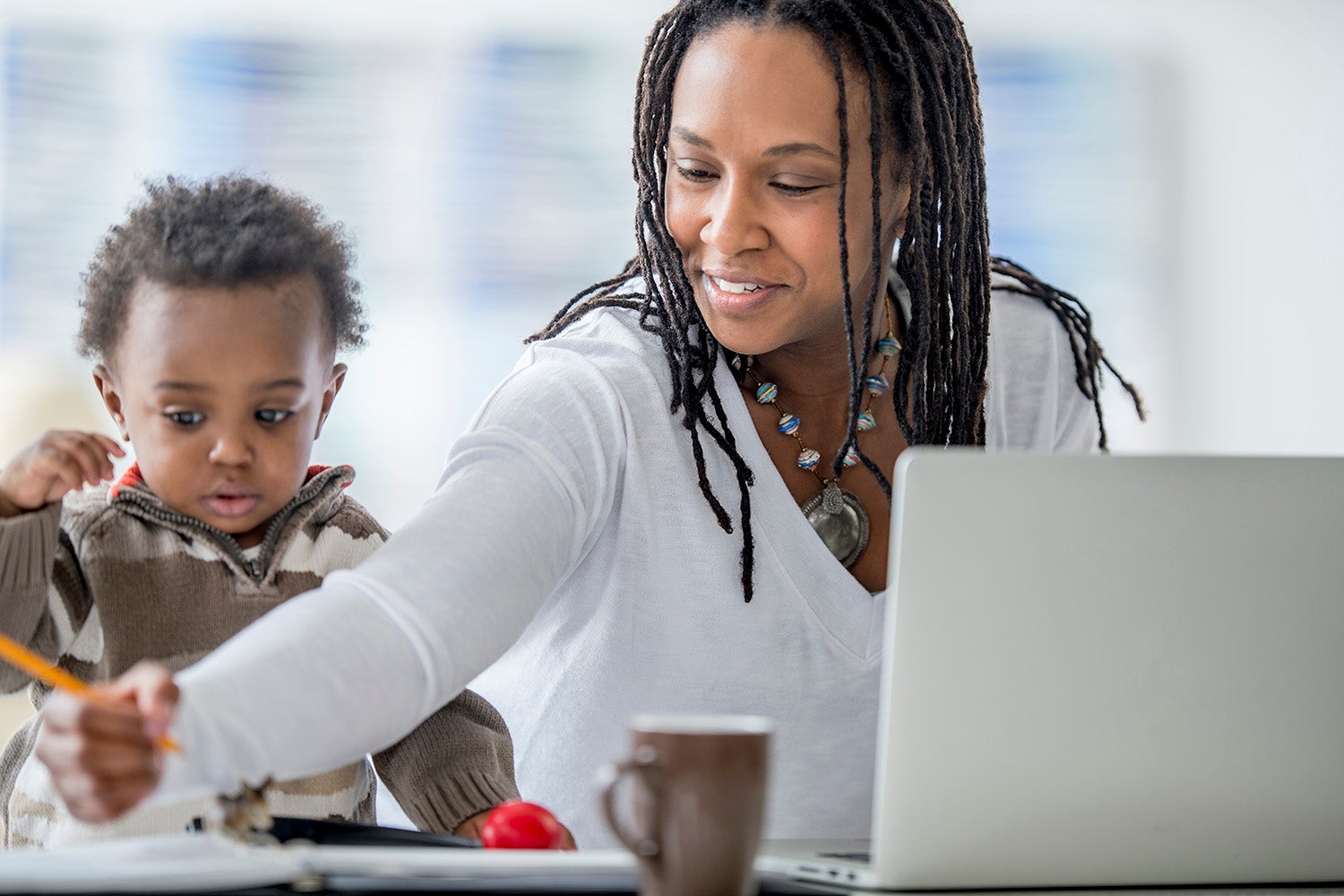 A woman of African descent and her baby boy are at home. The mother is working on a laptop computer. She is being distracted by her son, who was playing with a pencil.