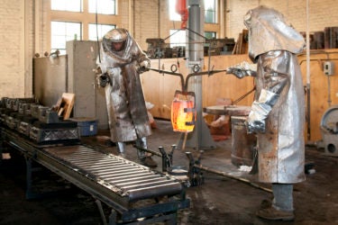Two people in full-body protective gear moving a container of molten metal toward molds