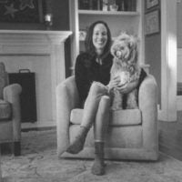 Amy Zegart smiling, sitting in a chair with her dog, whose head is cocked to the side