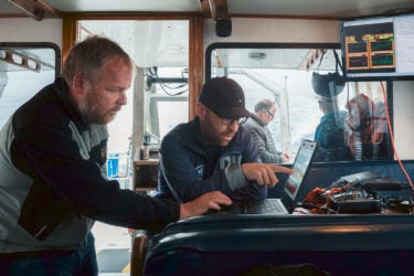 Two men inside a boat, looking at a computer screen