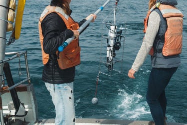 Two women near the back of a boat, pulling in a device using a hook