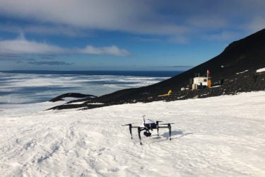 A drone on snow with a small building in the background and water in the distance