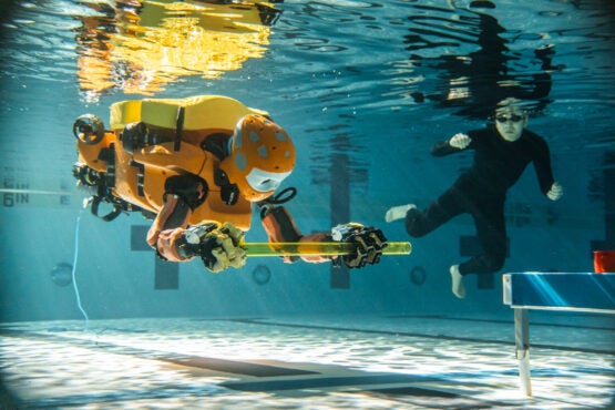 A humanoid robot and a person underwater in a swimming pool.  The robot holds a plastic rod with two hands.