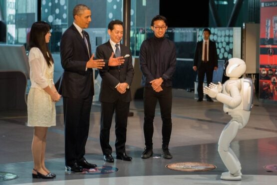 Four people facing a human-like robot. One person is Barack Obama. Obama and the robot have their hands raised in similar ways.